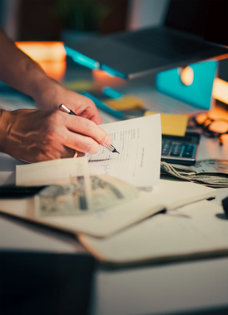 A hand signing financial documents on a desk with cash and a calculator, signifying finance management.